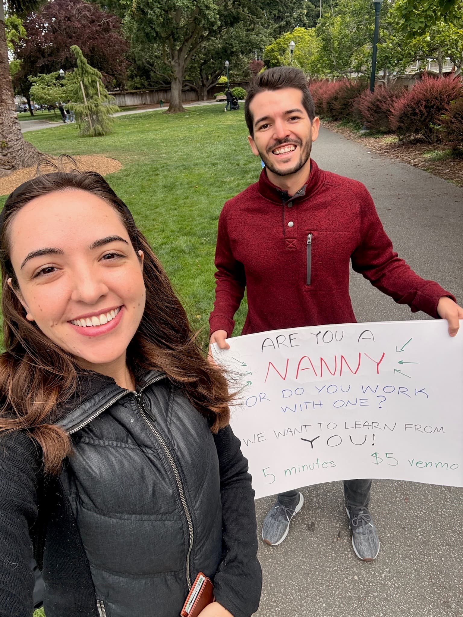 Jenna and Evan in a park holding a hand-made sign reading: are you a nanny, or do you work with one? We want to learn from you.